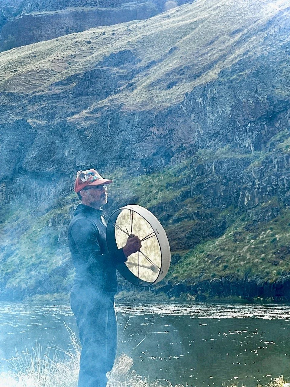 Gabe holding a frame drum beside the river.