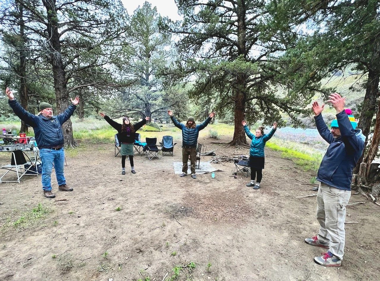 A group practising qi gong with arms raised in a riverside camp.